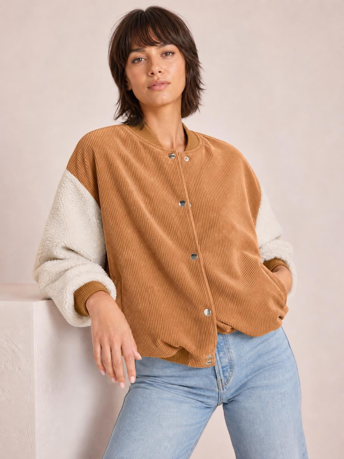 A model leans casually in a relaxed camel corduroy jacket with contrasting cream sherpa long sleeves, ribbed baseball collar, snap front, and roomy silhouette.