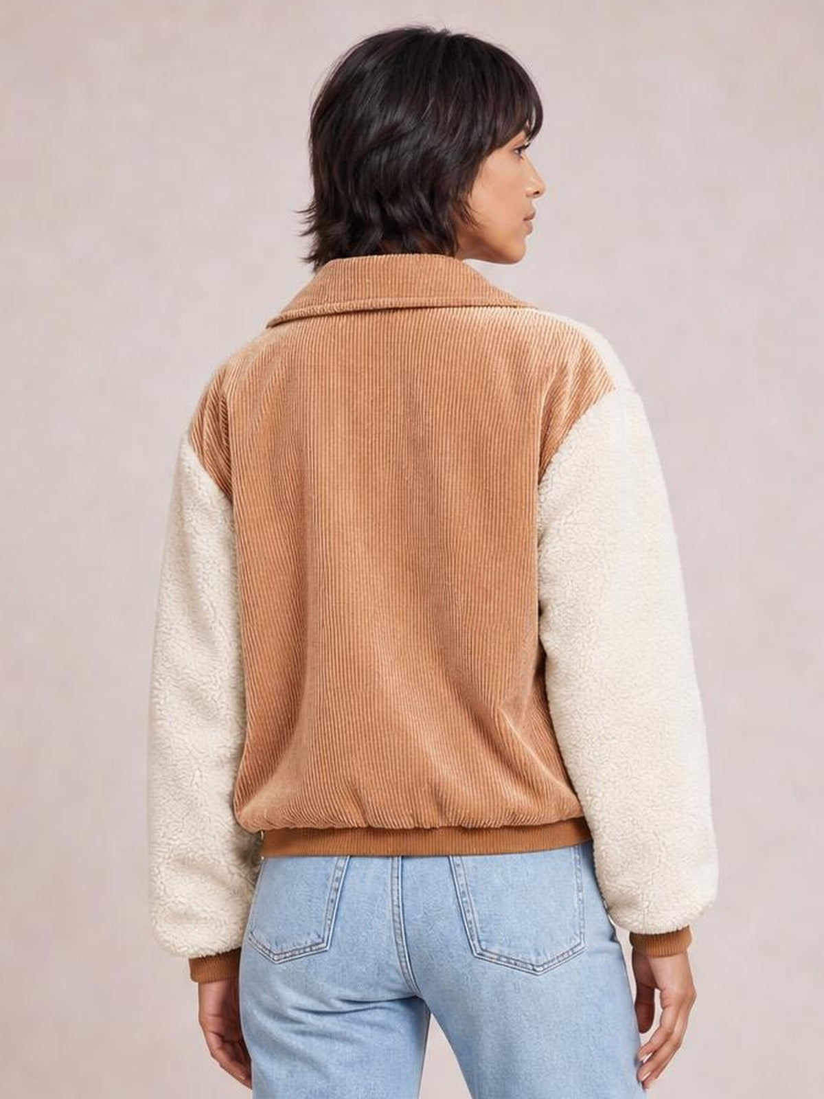 A woman models a relaxed contrast baseball jacket from the back, featuring a camel corduroy body, cream sherpa long sleeves, and ribbed collar, cuffs, and hem.