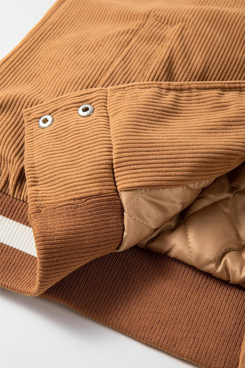 Close-up of a camel-brown ribbed jacket showing snap closures, a contrasting white stripe, ribbed cuff and hem, and quilted lining.