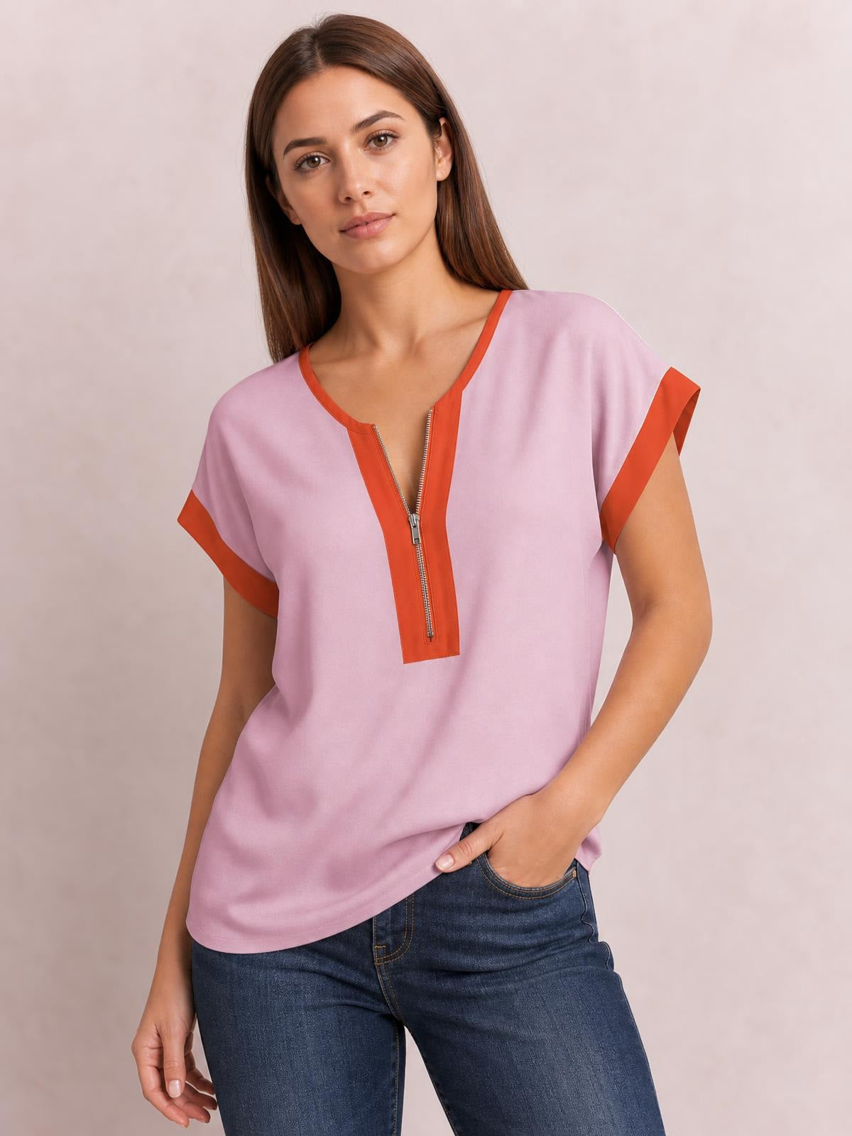 Model wearing a relaxed pink short-sleeve top with contrasting orange trim, a half-zip neckline, and curved hem, posing with one hand in her jeans pocket.
