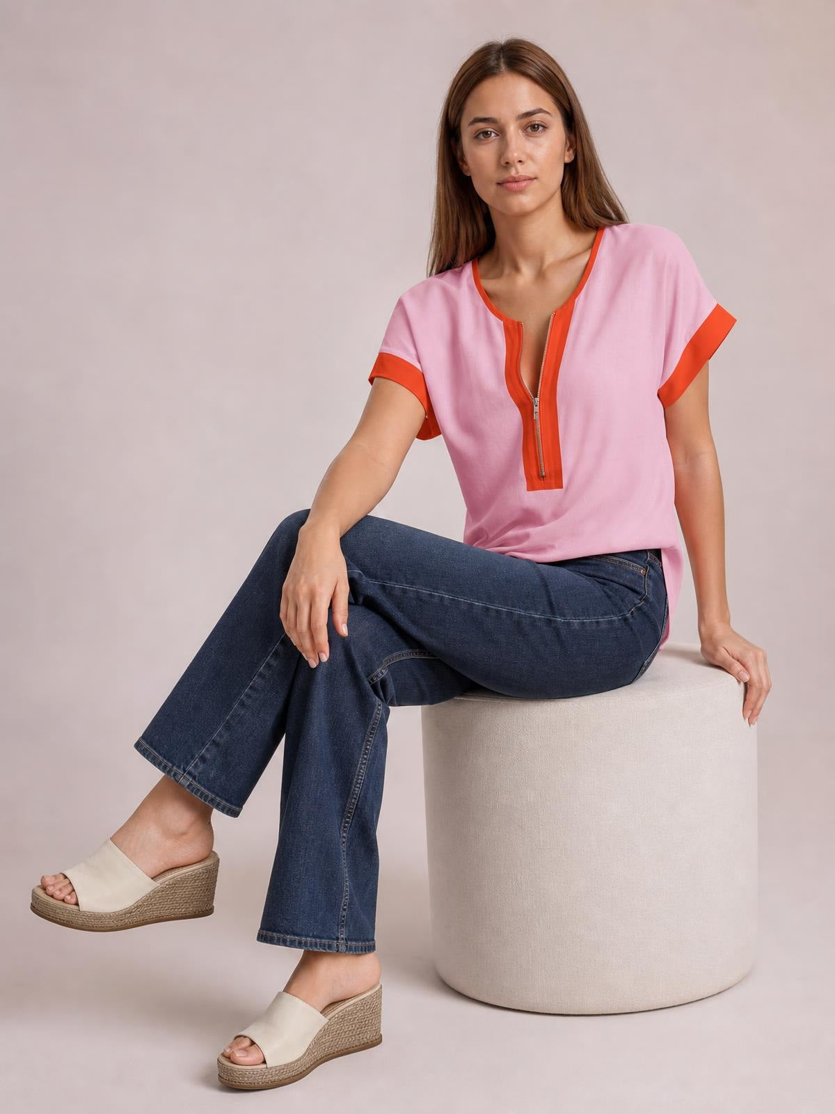 A seated model wears a pink contrast half-zip short-sleeve top with orange trim, a relaxed silhouette, and a partially open neckline.