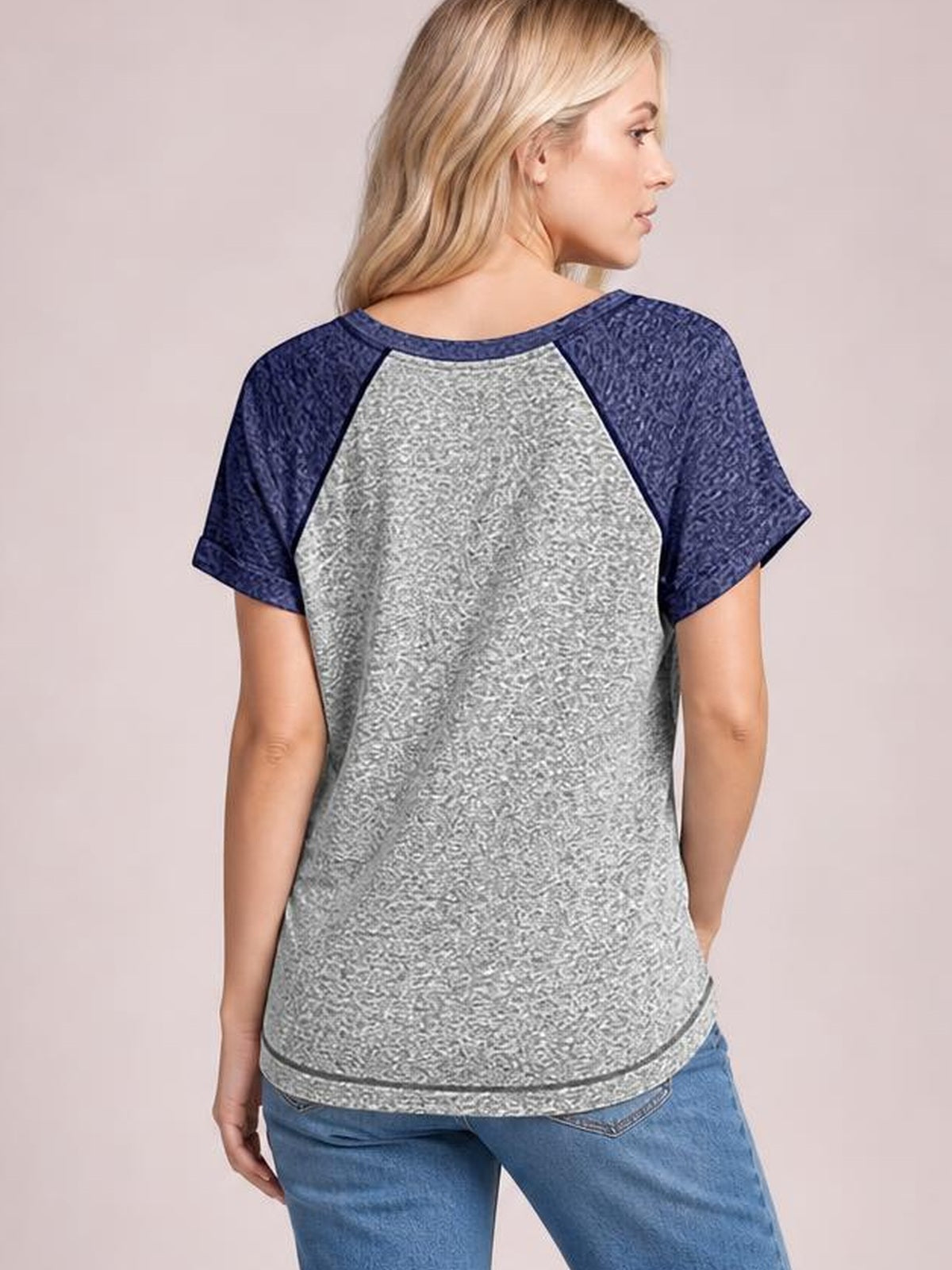 A model faces away in a gray contrast T-shirt with navy short raglan sleeves, a relaxed silhouette, curved hem, and textured panel construction.