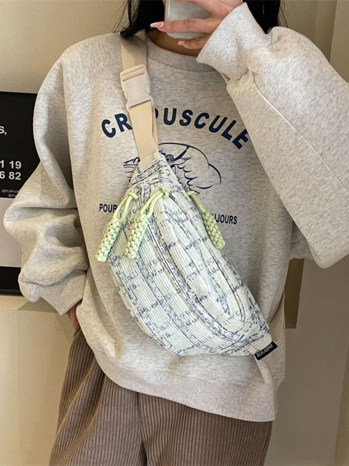 Model wears a softly curved polyester crossbody bag with a cream and navy contrast plaid pattern, wide strap, and playful lime cord accents.