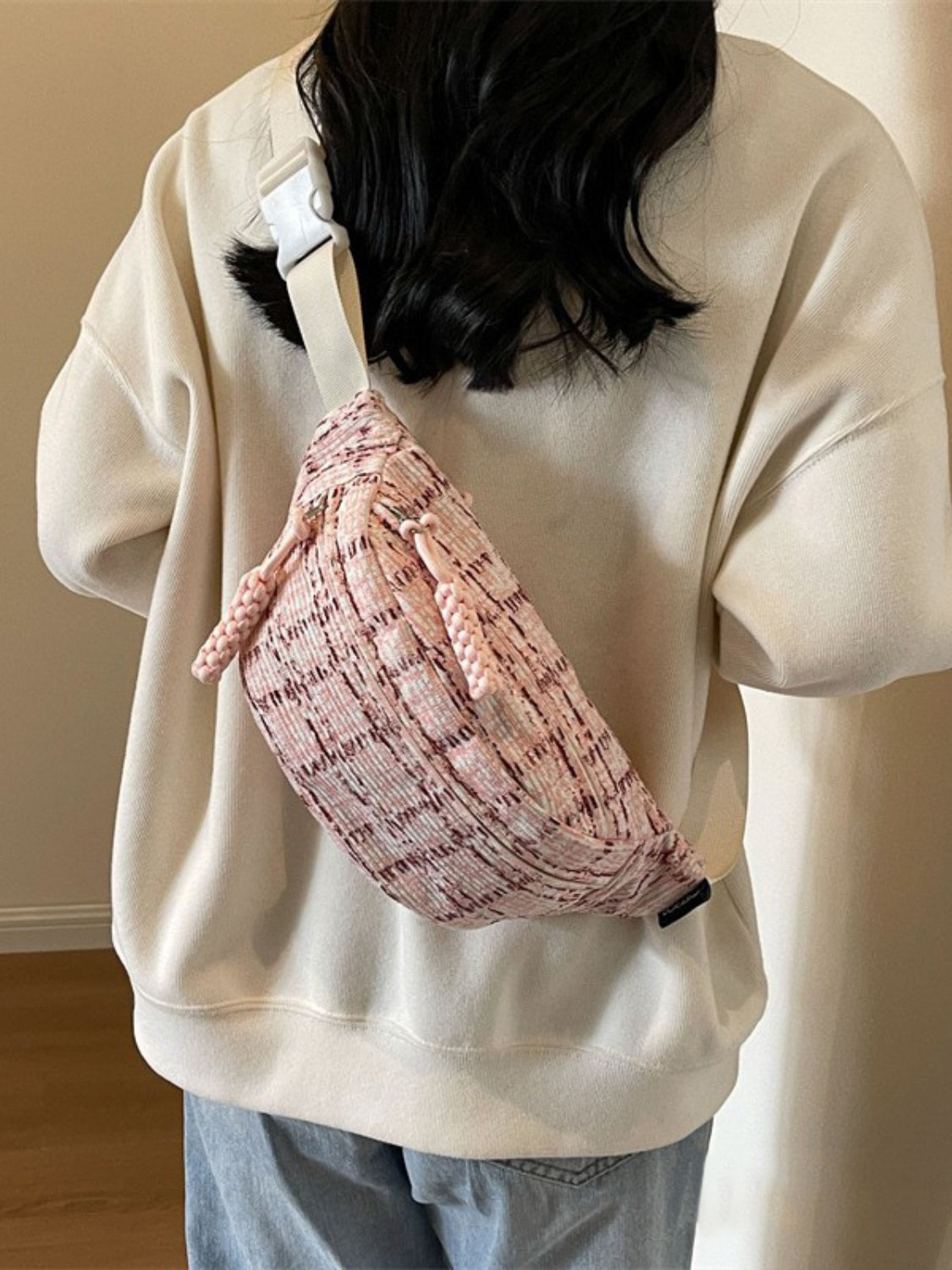 Model wearing a pink contrast plaid polyester crossbody bag with a softly curved silhouette, long strap, and visible cord accents over a cream sweatshirt.