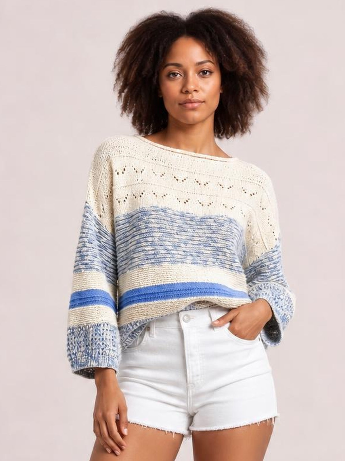 Model poses in a cream and blue contrast round-neck sweater with long sleeves, open-knit detailing, horizontal stripes, and a relaxed cropped silhouette.