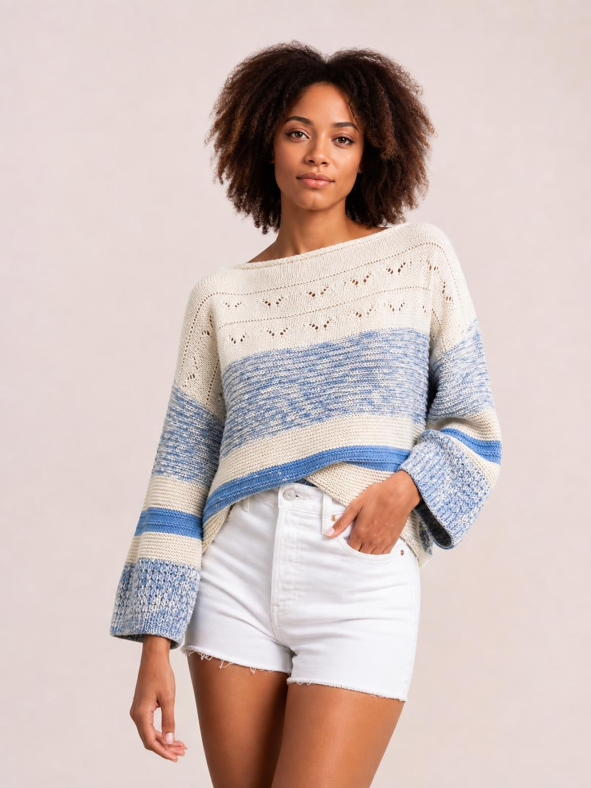 Model wearing a cream and blue contrast round neck long sleeve sweater with a relaxed silhouette, textured knit bands, and one hand tucked into white shorts.