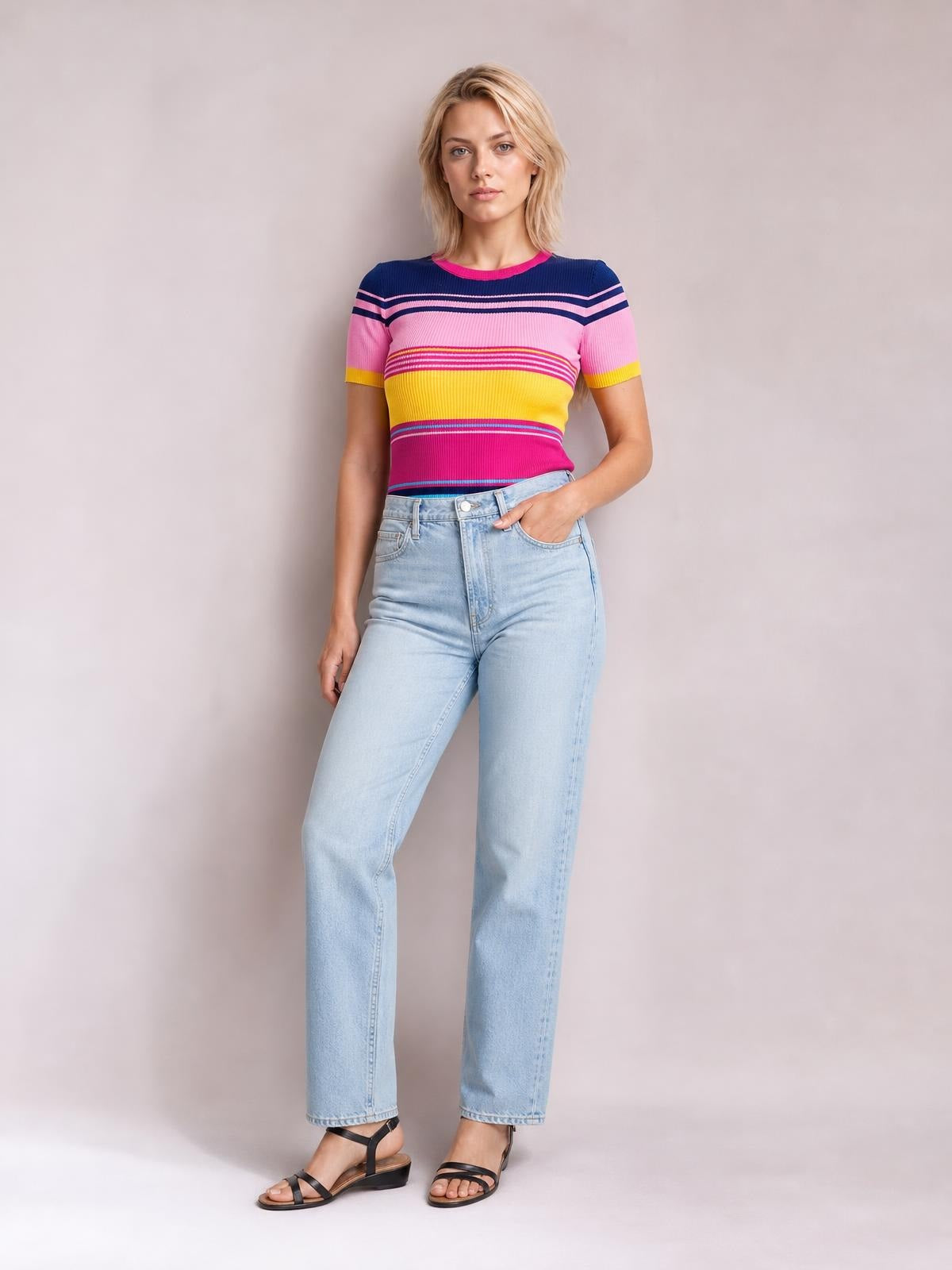 Model stands in light-wash jeans wearing a colorful contrast round-neck short-sleeve sweater with pink, yellow, and navy stripes, one hand in her pocket.