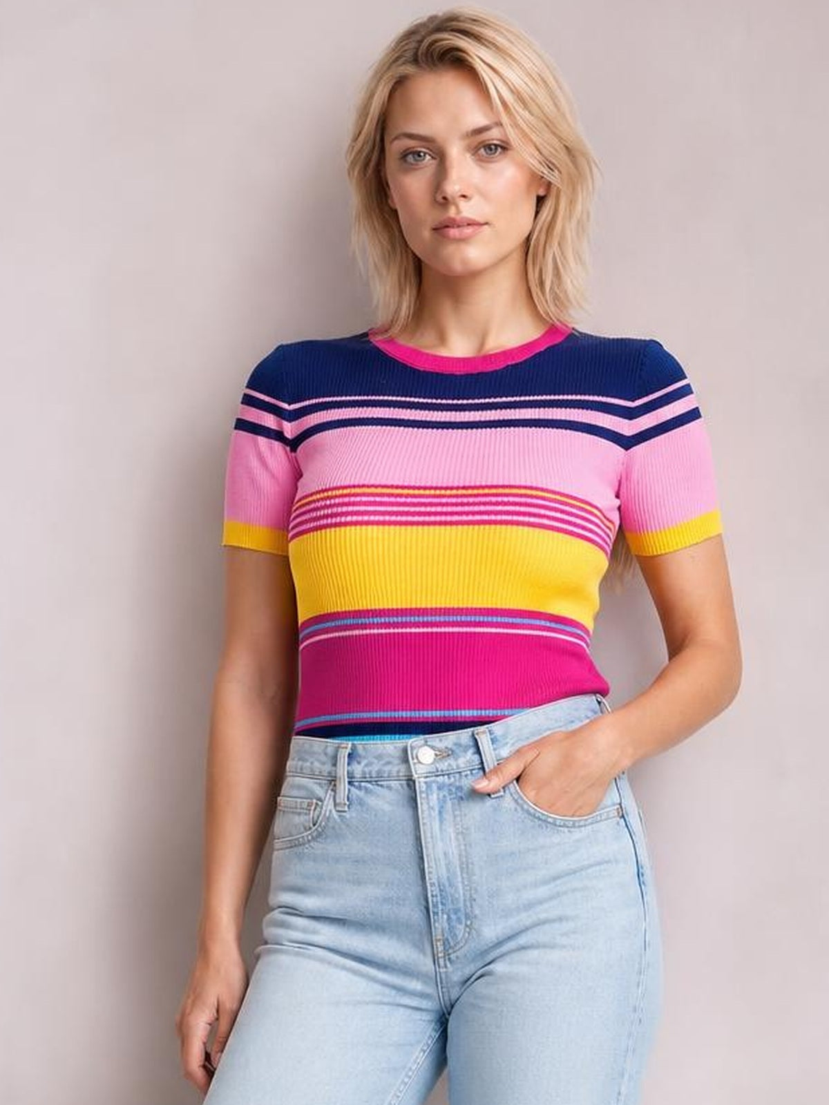 Model wearing a fitted contrast round neck short sleeve sweater with ribbed knit construction, bold navy, pink, yellow and magenta stripes, hand in jeans pocket.