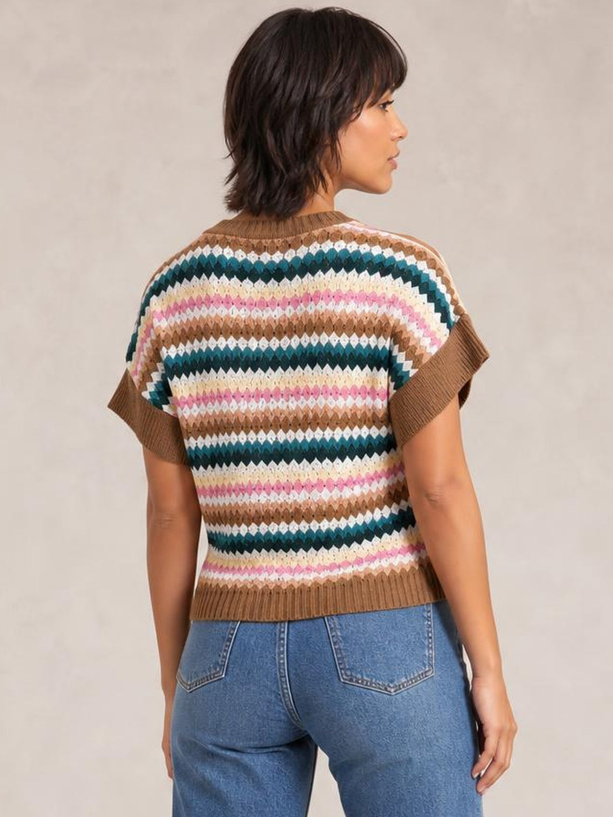 A model faces away wearing a multicolor contrast round-neck short-sleeve sweater with horizontal patterned knit, brown ribbed trim, and a relaxed silhouette.