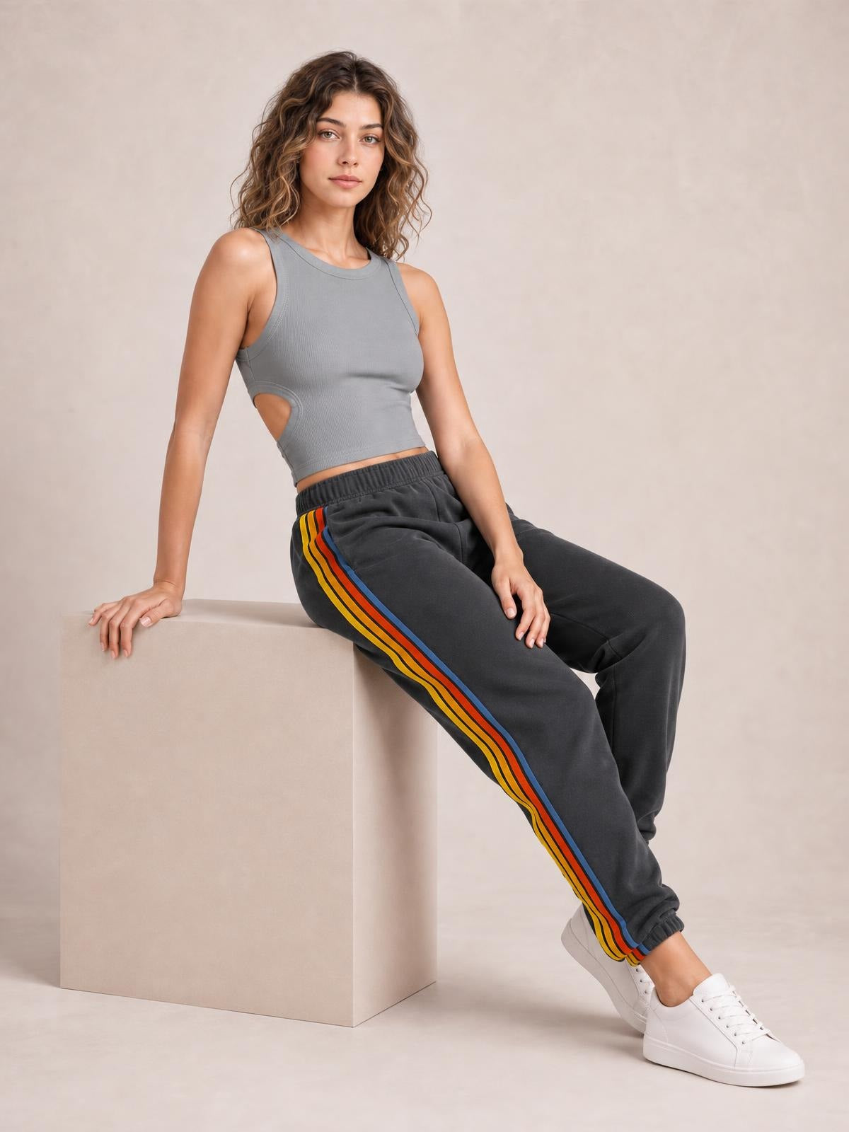 A model leans seated in dark active pants with an elastic waist, relaxed jogger silhouette, colorful contrast side stripes, and elastic ankle cuffs.