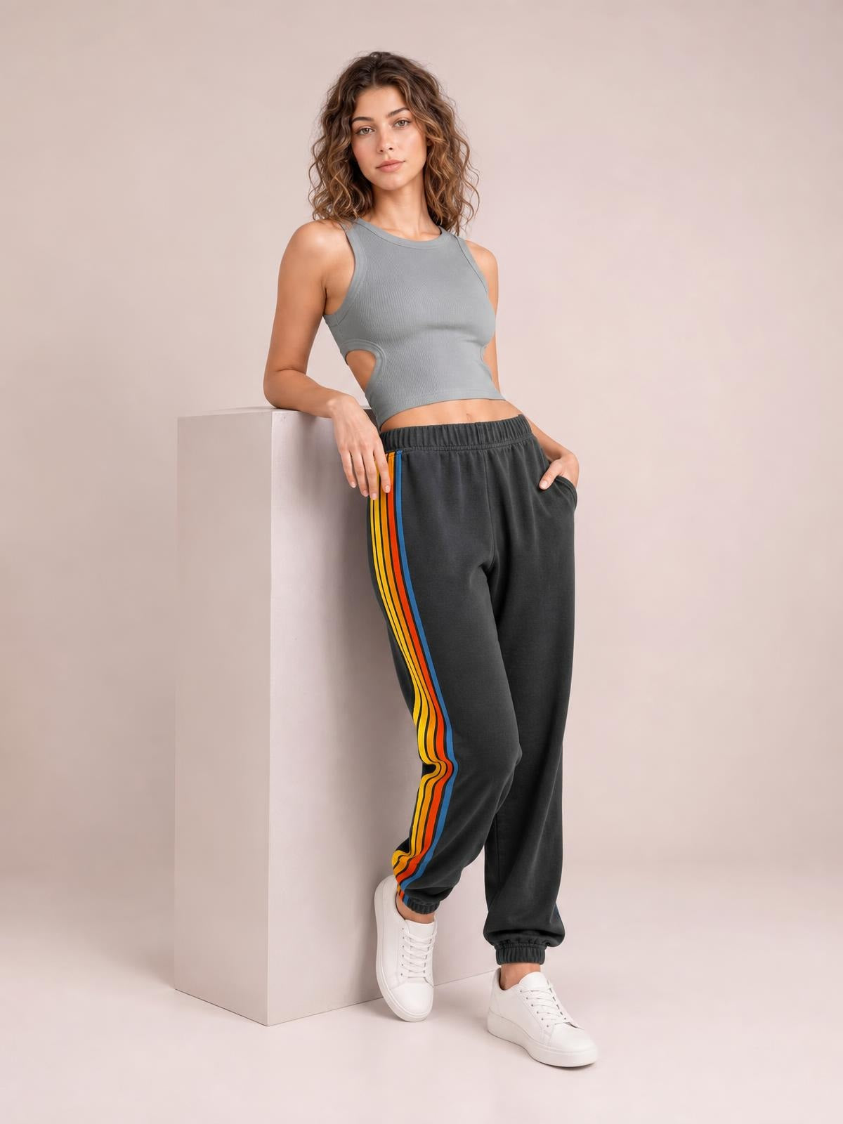 A model leans against a pedestal wearing dark gray active pants with a gathered elastic waist, colorful contrast side stripes, and a relaxed tapered silhouette.