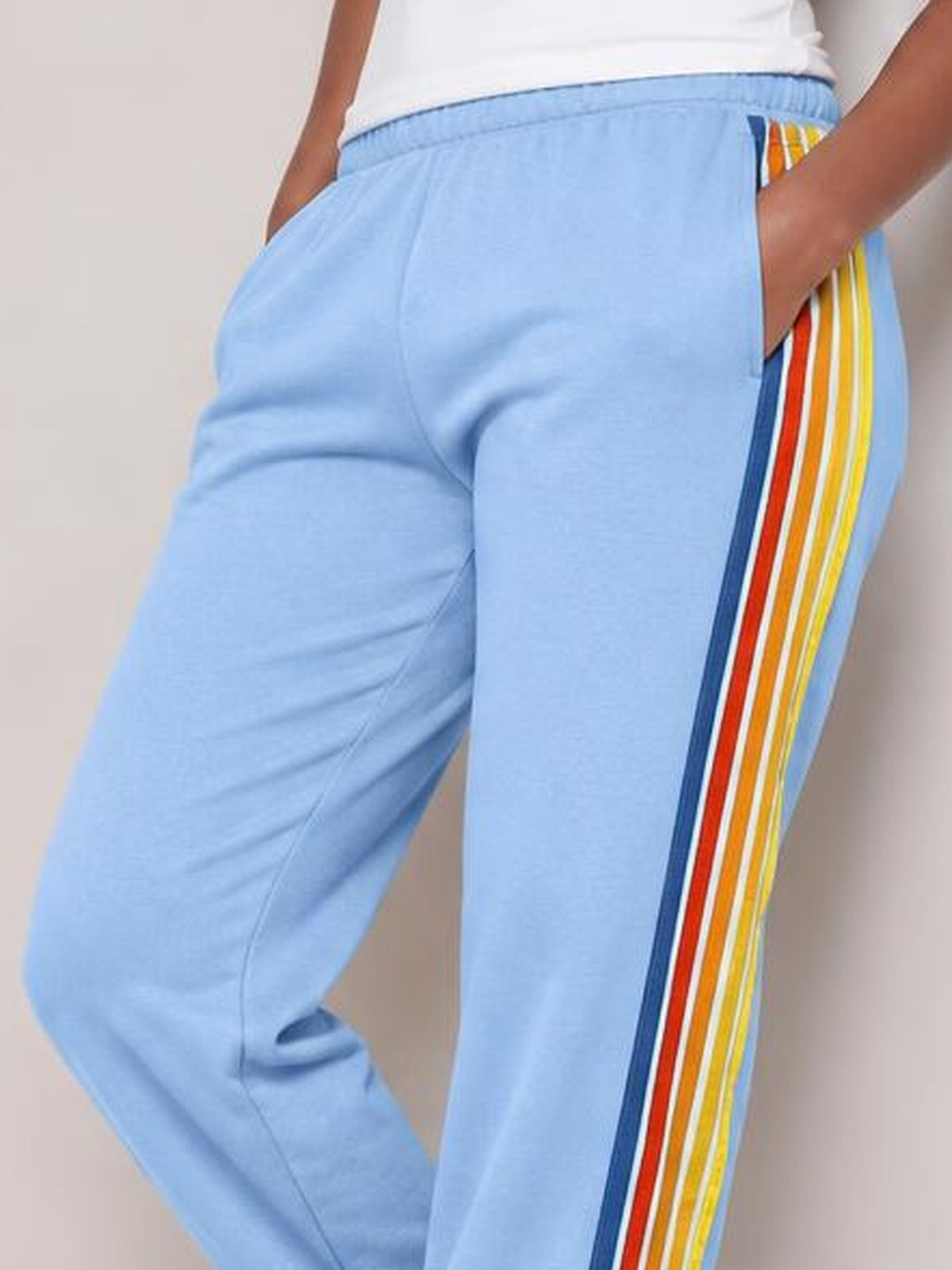 A model poses with hands in the pockets of light blue active pants featuring an elastic waist, relaxed fit, and bold multicolor contrast side stripes.