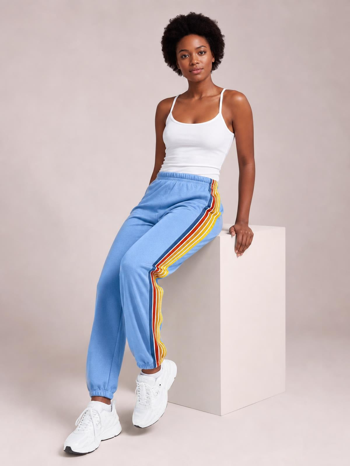 A model sits wearing light blue active pants with an elastic waist, multicolor contrast side stripes, a relaxed jogger silhouette, and cuffed ankles.
