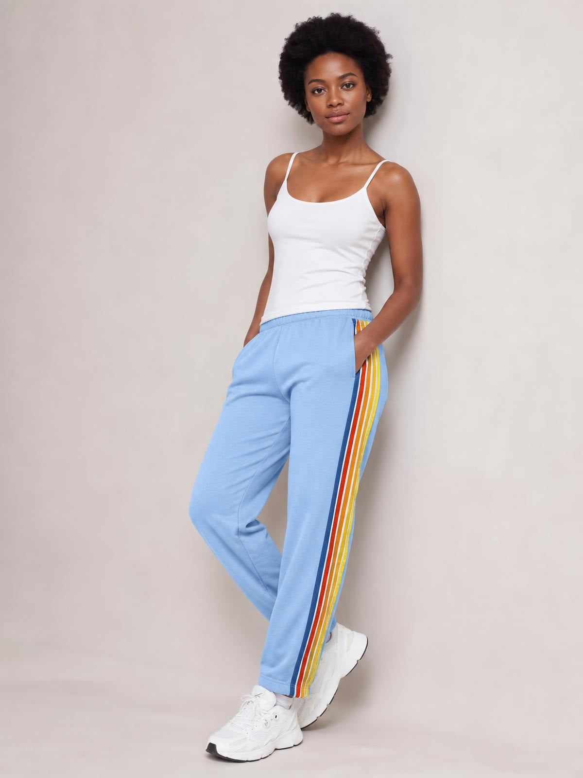 Model poses in light blue active pants with an elastic waist, relaxed straight-leg silhouette, and contrasting multicolor side stripes, with hands in pockets.