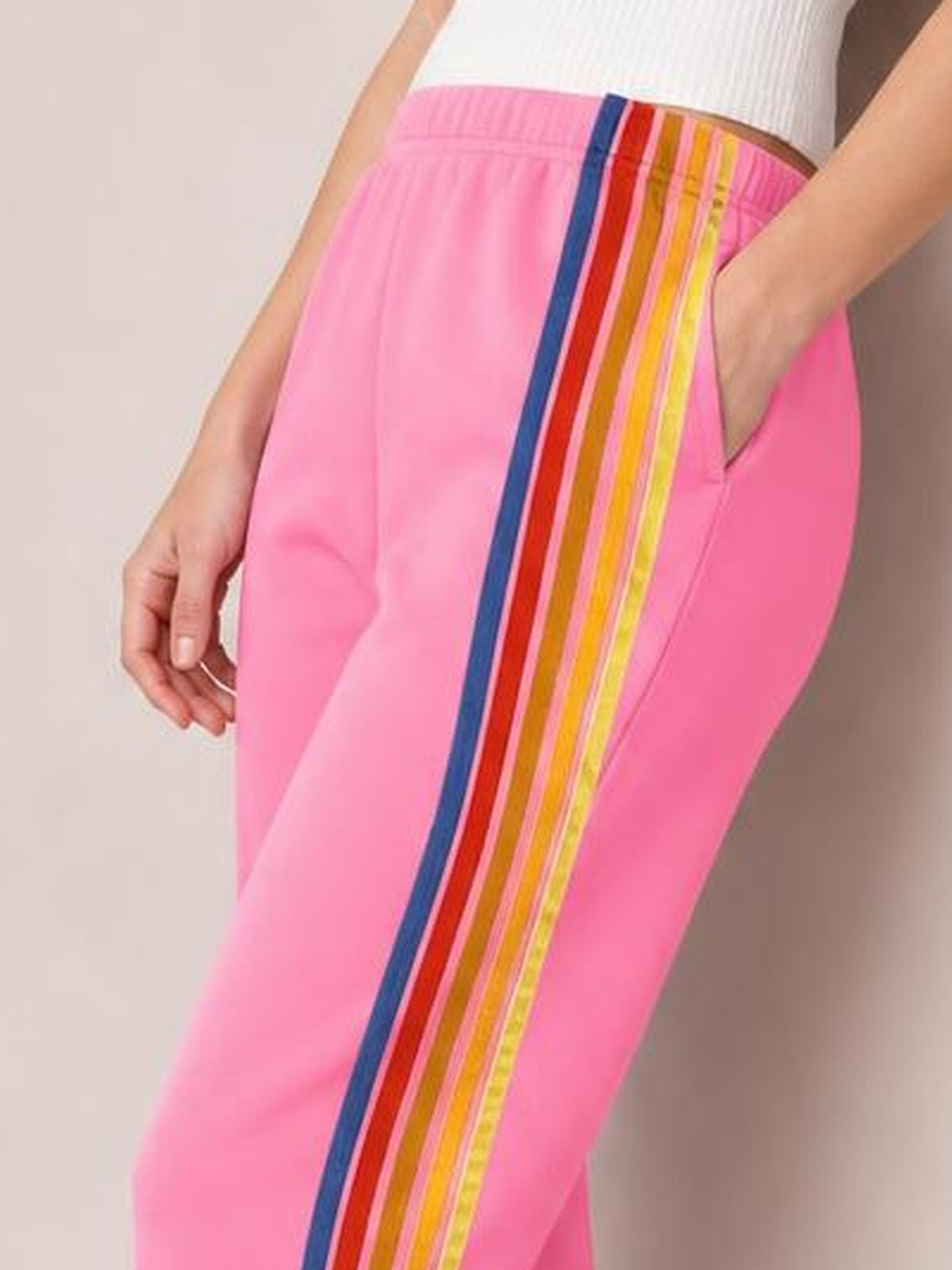 Model shown close up wearing bright pink active pants with an elastic waist, side pockets, and bold multicolor contrast stripes in a relaxed straight-leg silhouette.