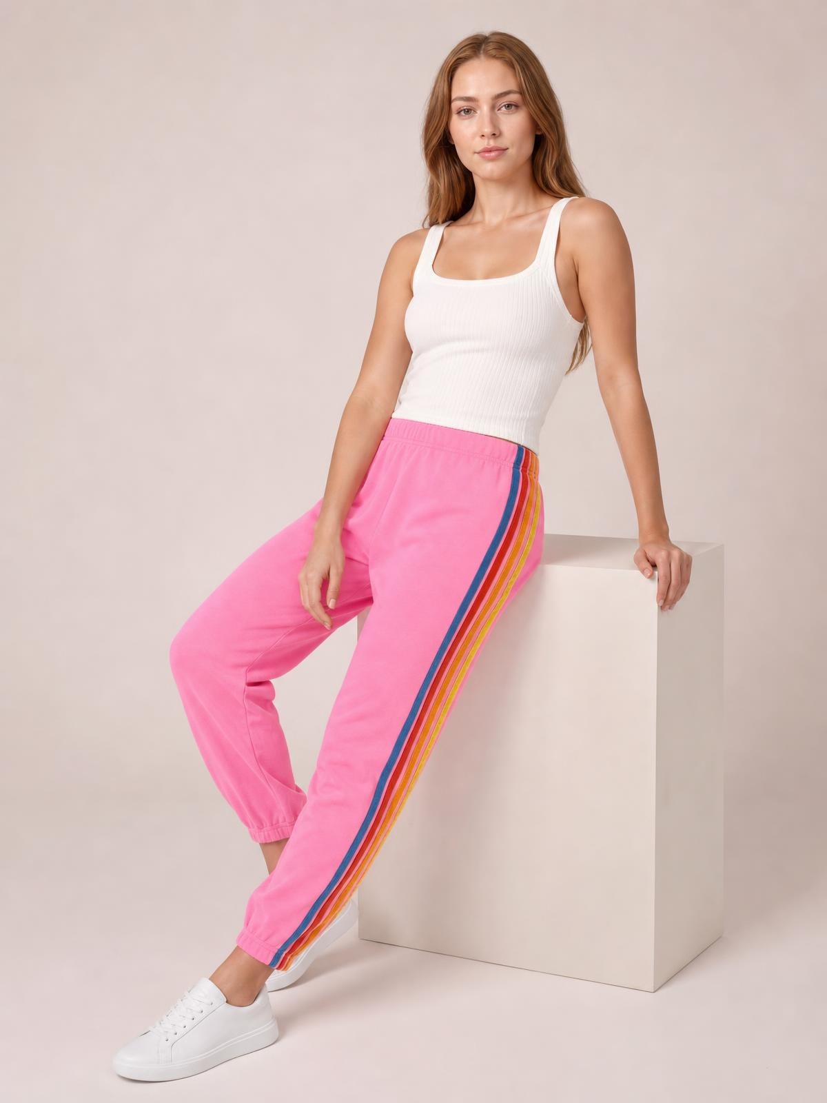 A model wears bright pink active pants with an elastic waist, multicolor contrast side stripes, a relaxed tapered silhouette, and elastic cuffs while seated.