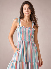 Model poses with one hand in a pocket, wearing a colorful contrast striped square-neck cami dress with a smocked bodice, tied straps, frill trim, and tiered skirt.