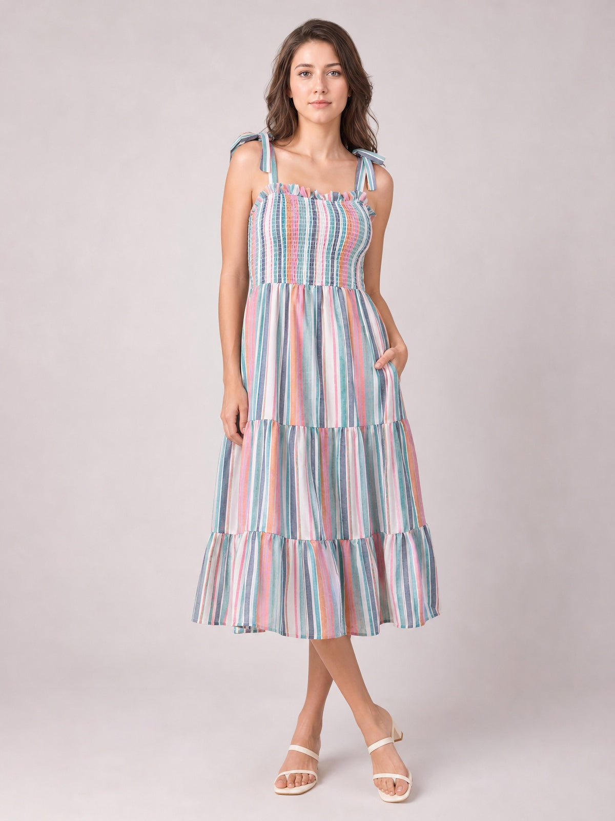 Model poses in a colorful contrast striped cami dress with tied shoulder straps, a smocked ruffled bodice, pocketed tiered midi skirt, and crossed legs.