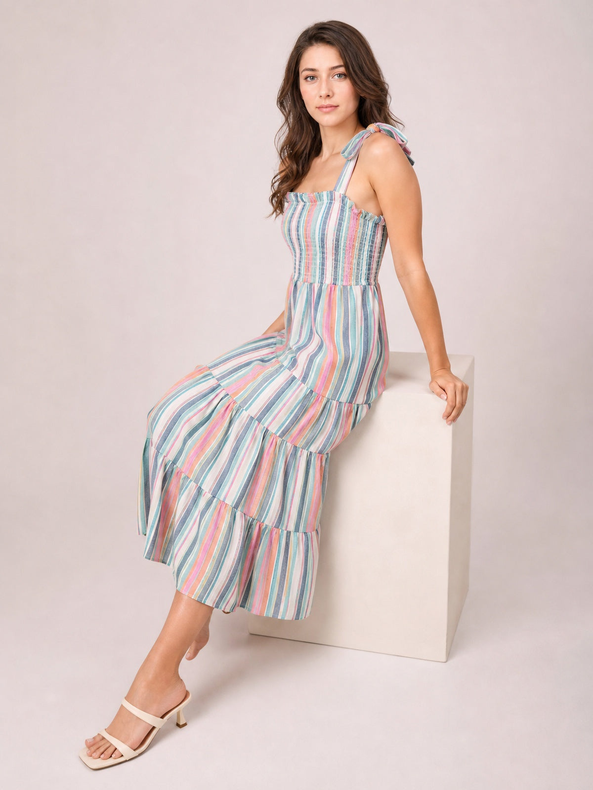 A seated model wears a colorful contrast-striped square neck cami dress with tied straps, a smocked bodice, and a flowing tiered midi skirt.