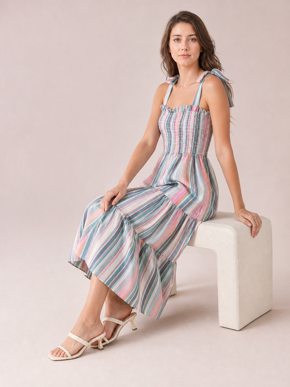 A seated model wears a colorful vertically striped cami dress with a square ruffled neckline, tied shoulder straps, smocked bodice, and flowing tiered midi skirt.