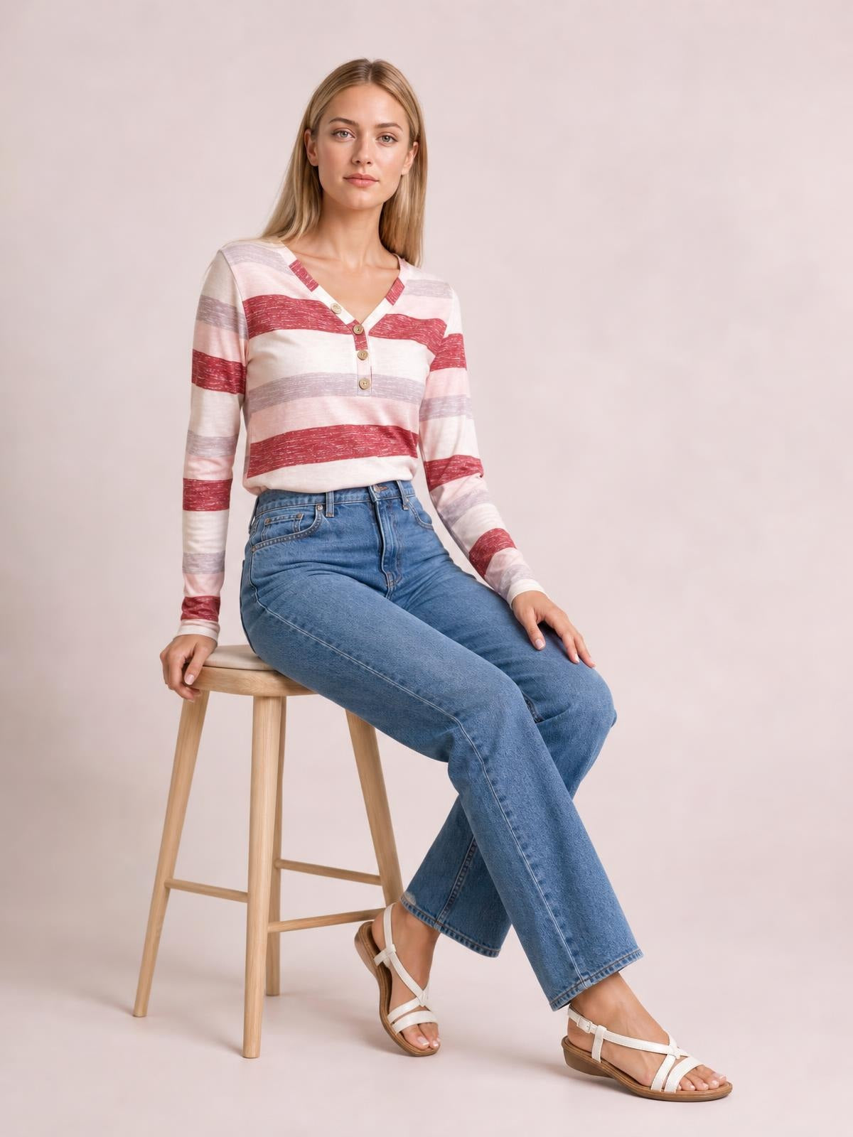Model seated with one hand resting beside her, wearing a cream contrast striped V-neck long-sleeve T-shirt with red and gray bands and a button placket.
