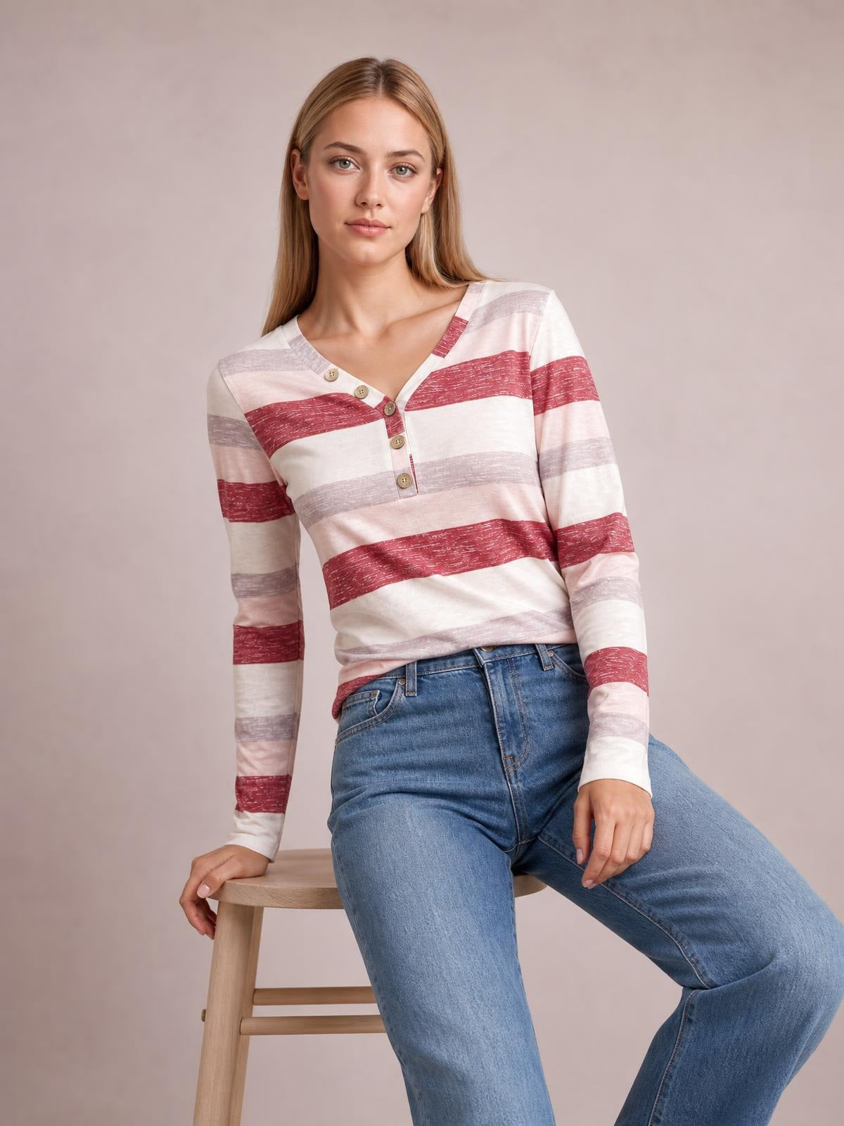 Model seated in blue jeans wearing a pink, burgundy, and gray contrast striped V-neck long sleeve T-shirt with a button placket and relaxed silhouette.