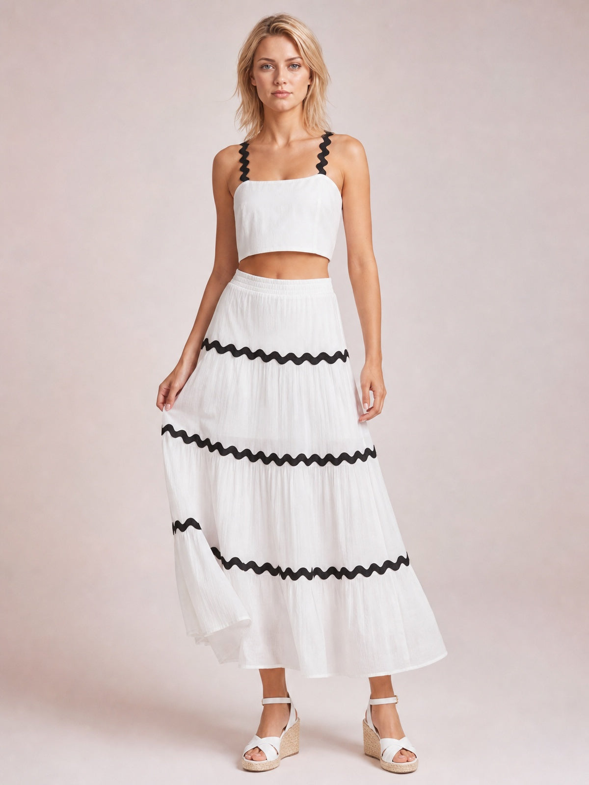 Model stands in a white cami and long skirt set with black wavy contrast trim, straight neckline, cropped silhouette, and tiered flowing construction.