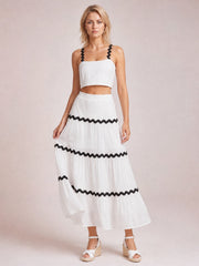 Model stands in a white cami and long skirt set with black wavy contrast trim, straight neckline, cropped silhouette, and tiered flowing construction.