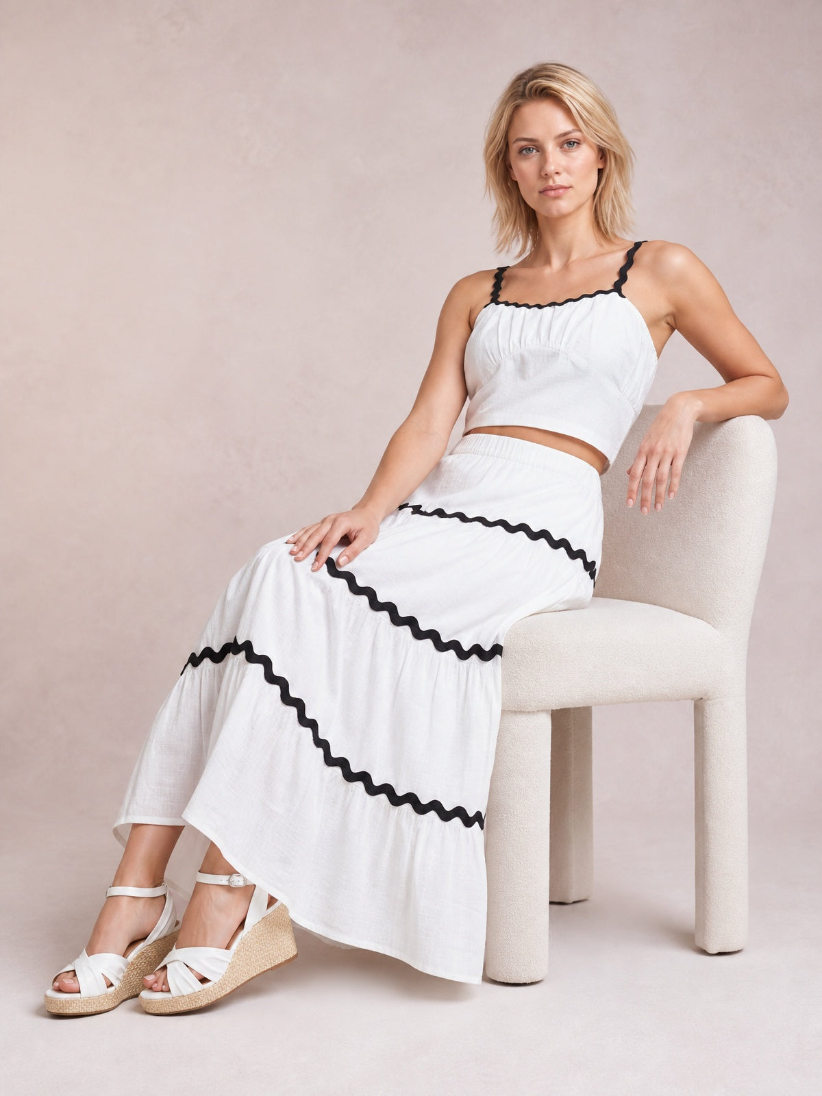 A seated model wears a white ruched cami and long tiered skirt set with black wavy contrast trim, a cropped silhouette, and slender straps.