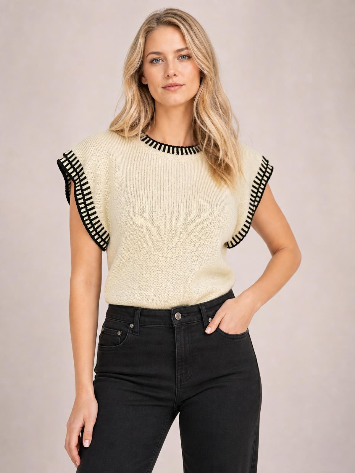 Model poses with one hand in her pocket wearing an ivory ribbed sweater with black contrast trim, cap sleeves, a crew neckline, and a relaxed straight silhouette.
