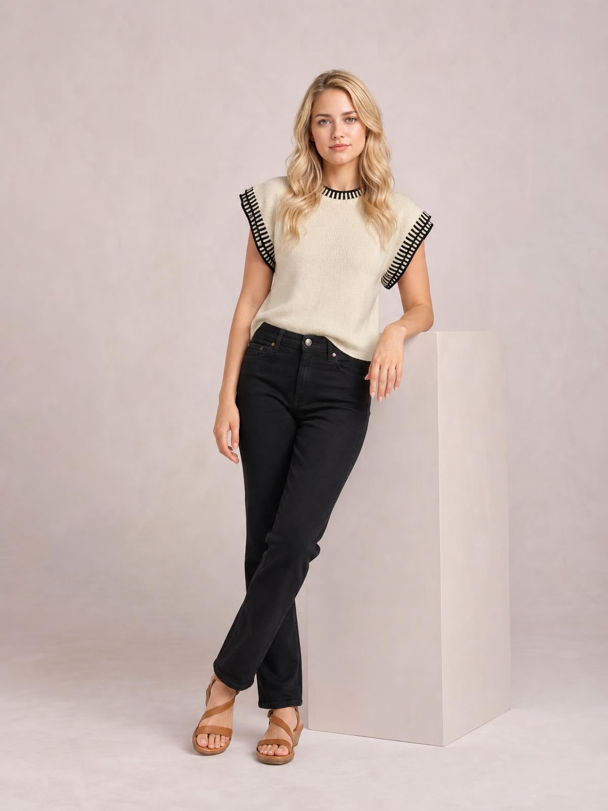 A model wears a cream ribbed sweater with cap sleeves, black contrast trim at the neckline and sleeve edges, and a relaxed straight silhouette while standing with crossed legs.