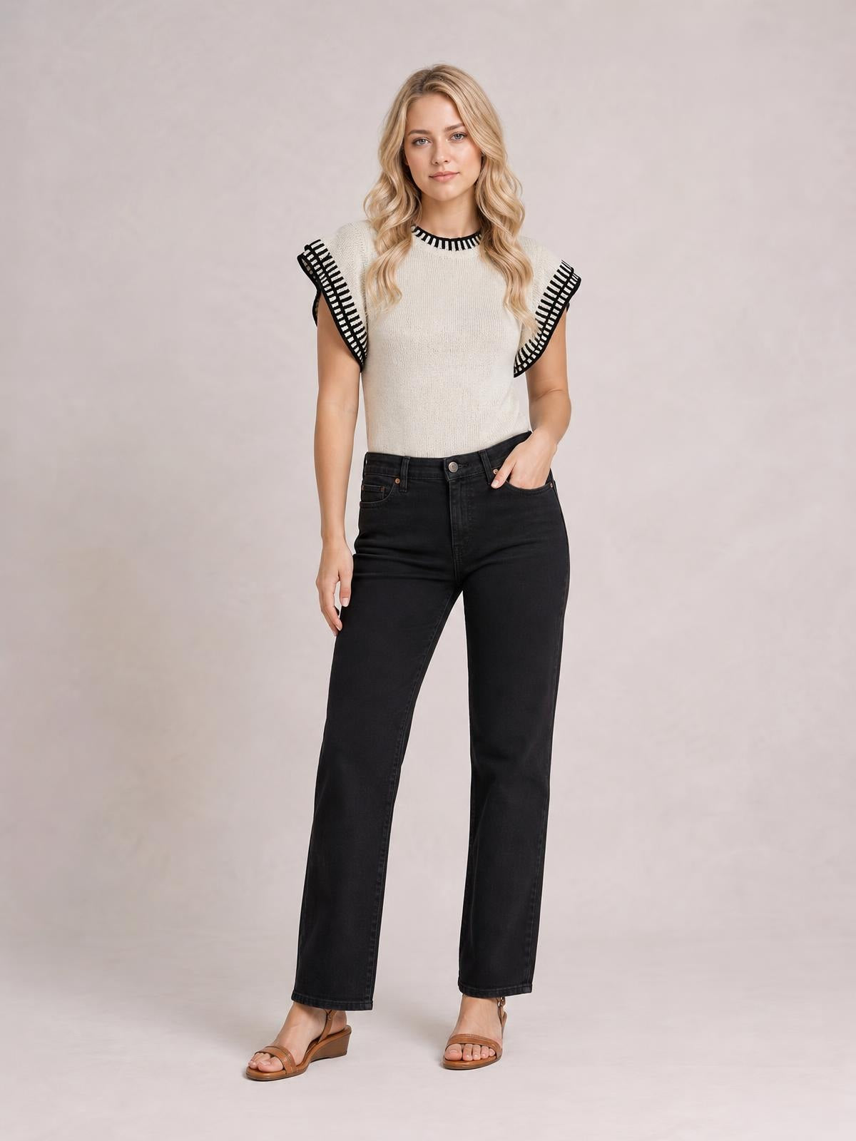 Model wears a cream ribbed contrast trim cap sleeve sweater with black ruffled edging, a fitted silhouette, and one hand resting in her jeans pocket.