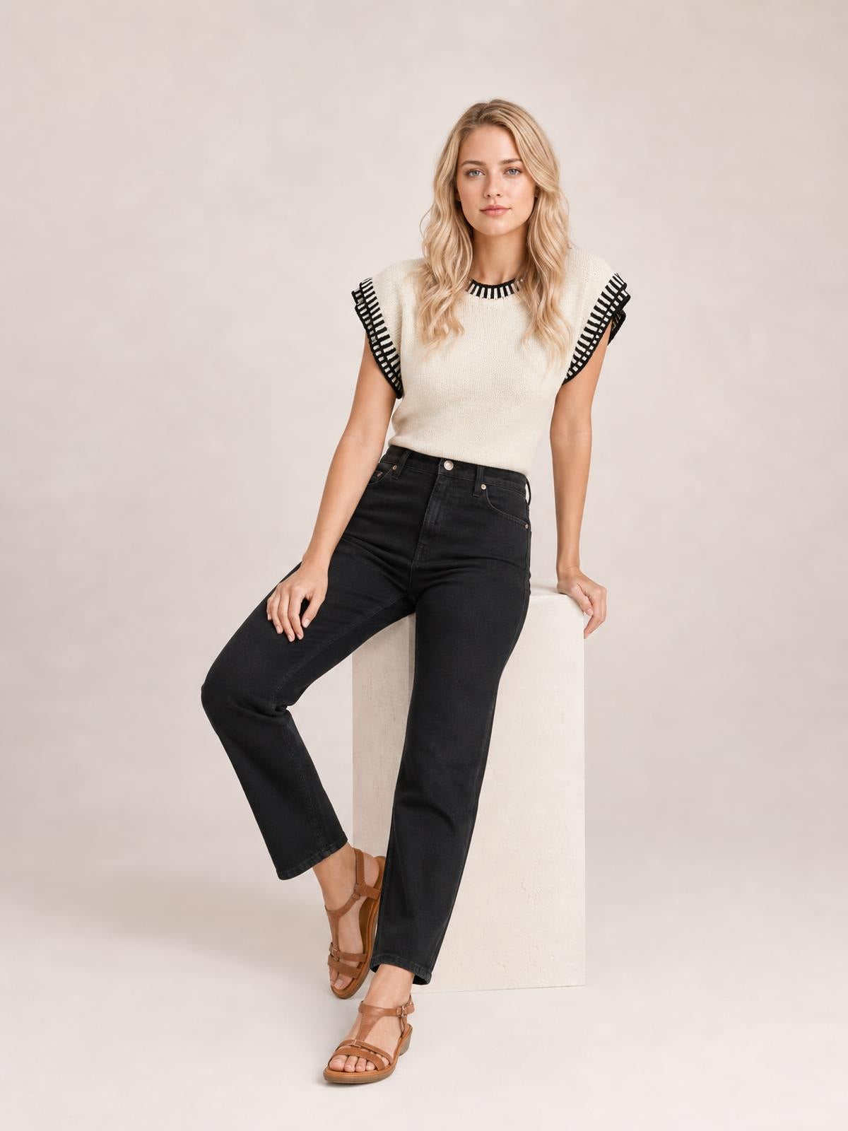 A woman models a cream ribbed sweater with contrasting black trim, cap sleeves, and a fitted silhouette while seated with one leg extended.