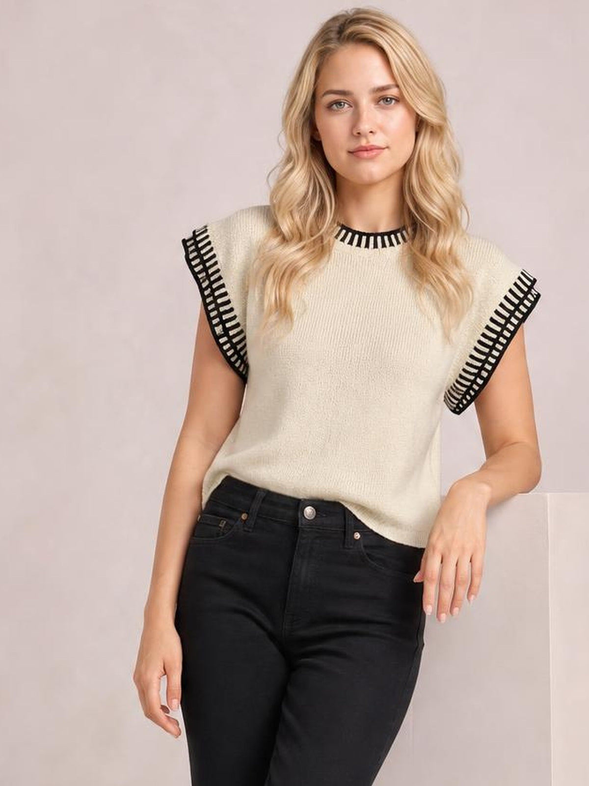 A model stands wearing a cream ribbed sweater with black contrast trim, cap sleeves, a round neckline, and a relaxed silhouette with a slightly curved hem.