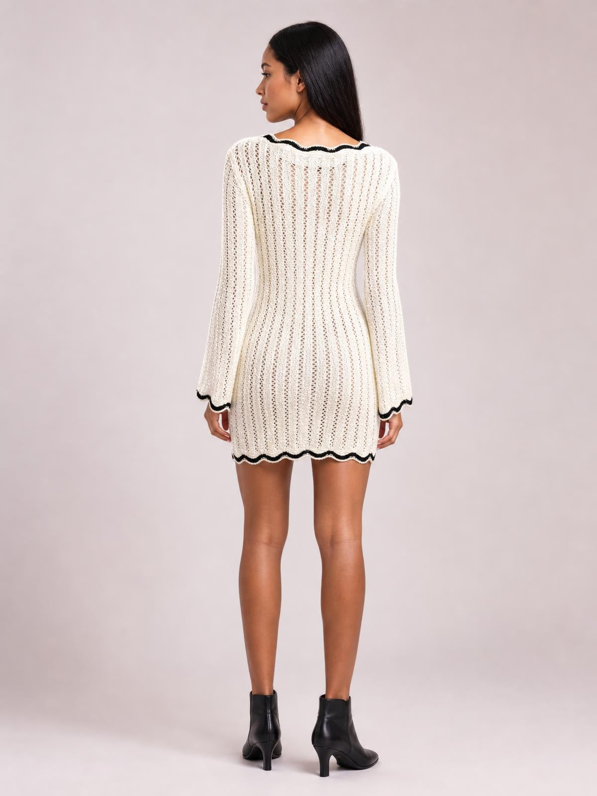 Model shown from the back in a cream crochet knit sweater dress with openwork vertical texture, fitted waist, long sleeves, and black contrast trim.