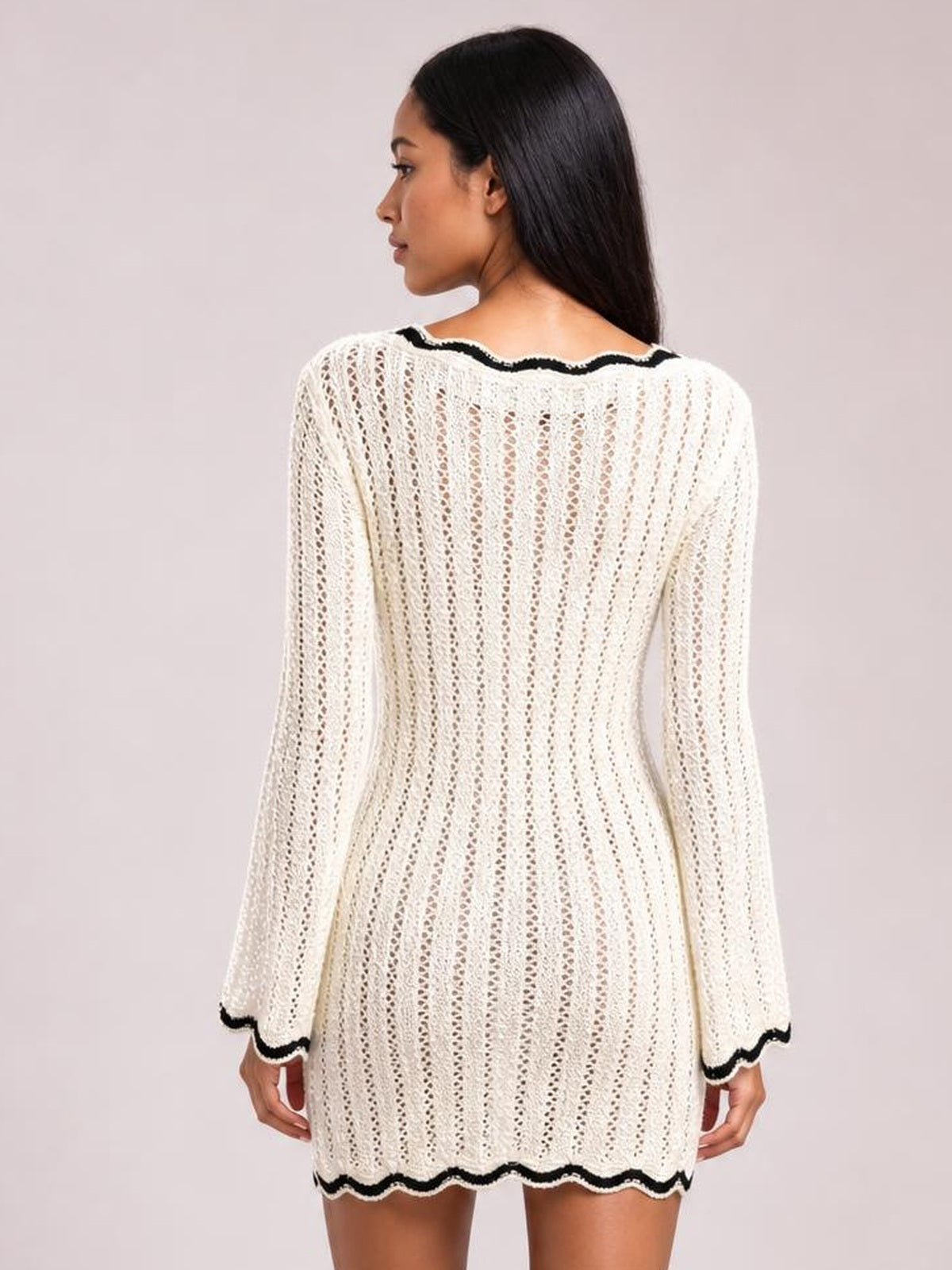 A model stands in a back view wearing a cream crochet knit sweater dress with openwork texture, long sleeves, and contrasting black scalloped trim at the neckline, cuffs, and hem.