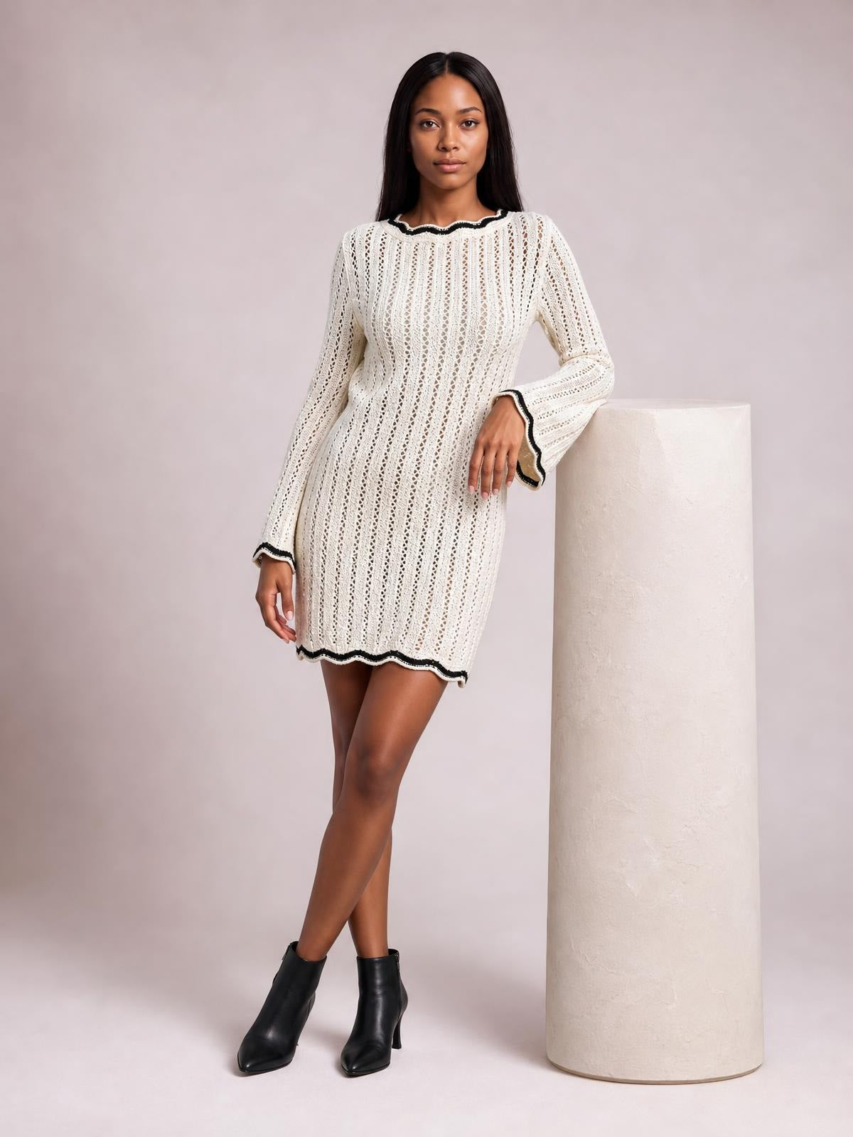 A model poses in a cream crochet knit sweater dress with openwork, long sleeves, a fitted mini silhouette, and black contrast trim at the neckline, cuffs, and hem.