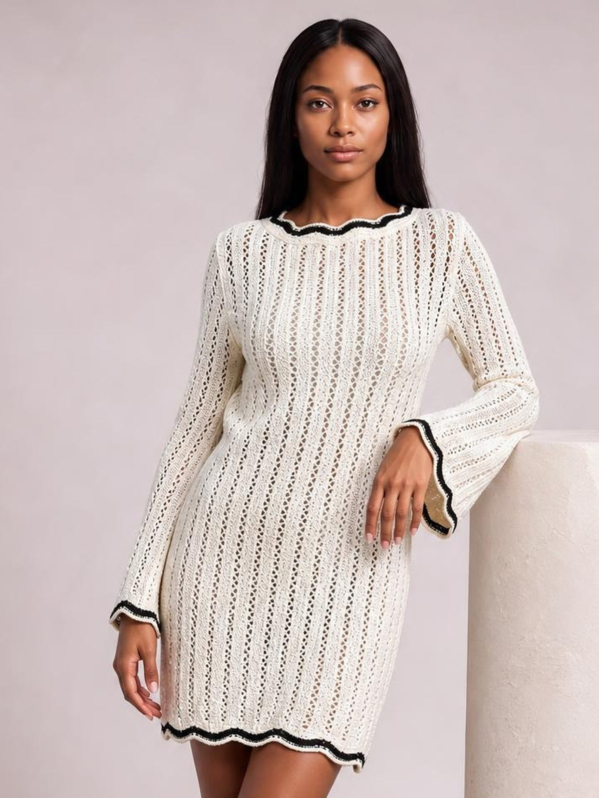 Model poses in a cream contrast-trim crochet knit sweater dress with openwork texture, long sleeves, a relaxed straight silhouette, and black edging.
