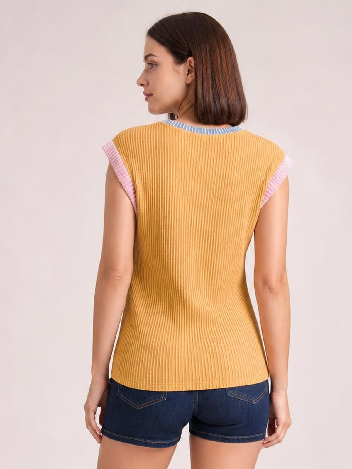 Model seen from behind wearing a mustard yellow ribbed knit sleeveless top with pink contrast armhole trim, blue neckline, and a fitted silhouette.