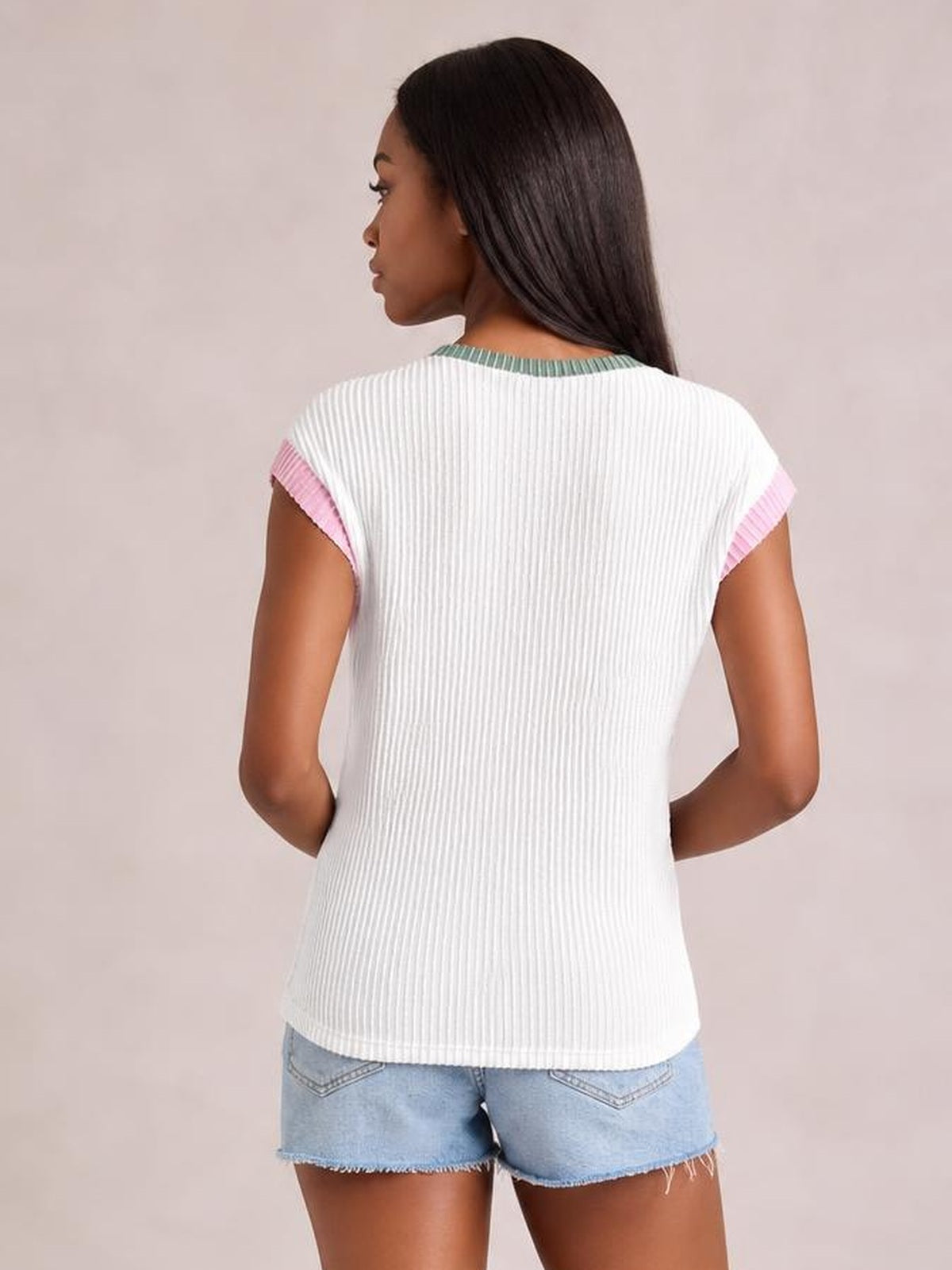 Model shown from behind wears a white ribbed knit top with pink sleeve trim, a green neckline, relaxed silhouette, and hands resting at her waist.