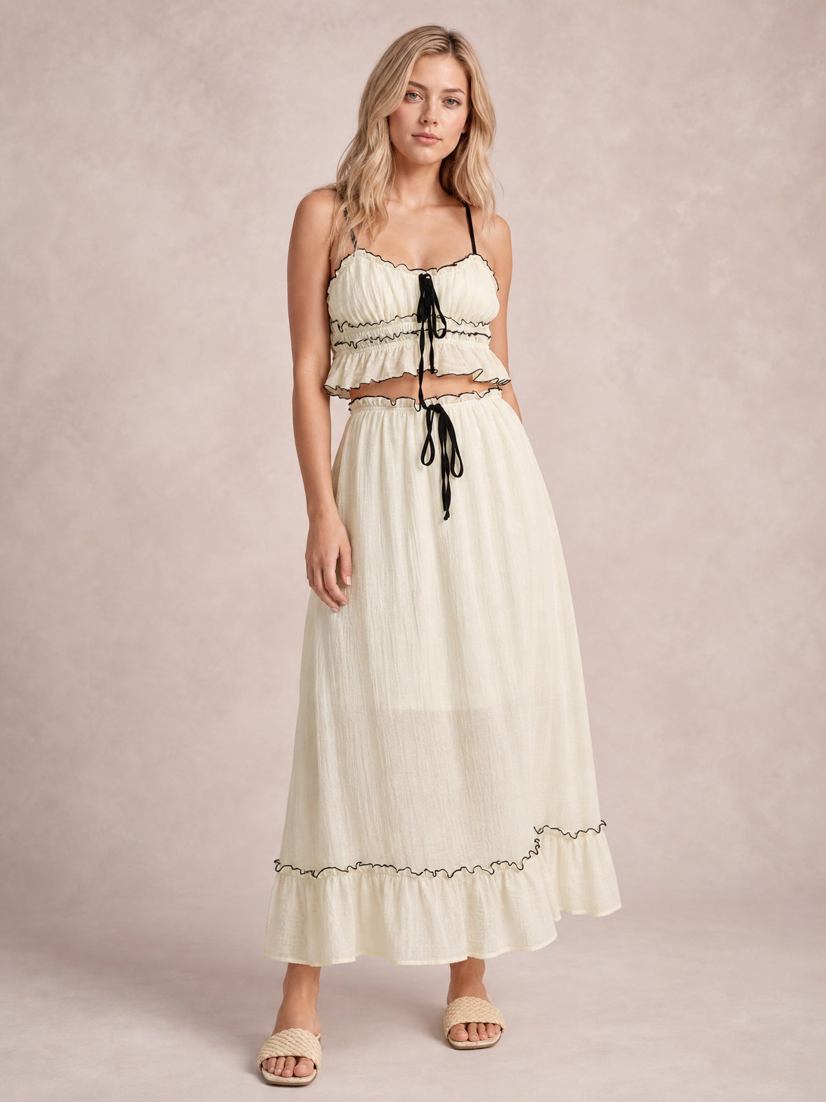A model stands in a cream cami and flowing midi skirt set with black contrast trim, smocked details, tied accents, delicate straps, and ruffled hems.