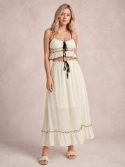 A model stands in a cream cami and flowing midi skirt set with black contrast trim, smocked details, tied accents, delicate straps, and ruffled hems.