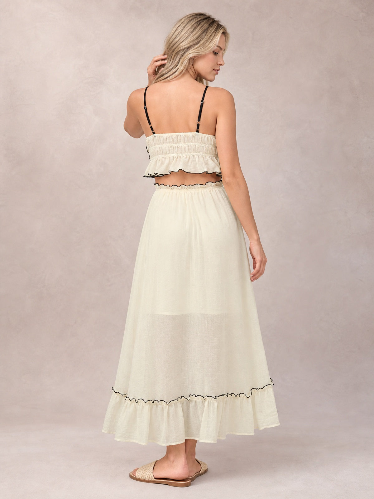 A model poses from behind in a cream cami and skirt set with thin straps, a smocked bodice, contrast-trimmed ruffles, and a flowing tiered midi skirt.