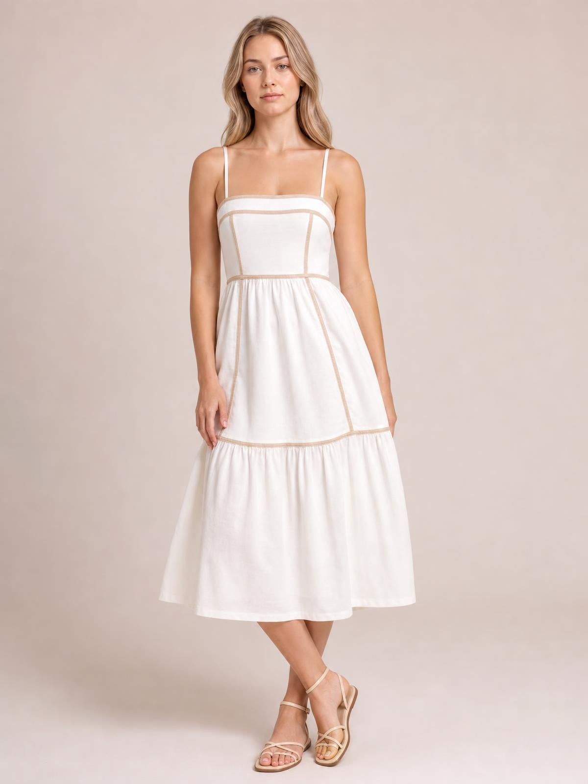 A model stands with crossed legs in a white square-neck midi cami dress featuring contrast trim, slender straps, a paneled bodice, and a gathered skirt.
