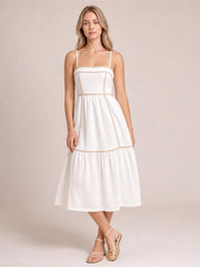 A model stands with crossed legs in a white square-neck midi cami dress featuring contrast trim, slender straps, a paneled bodice, and a gathered skirt.