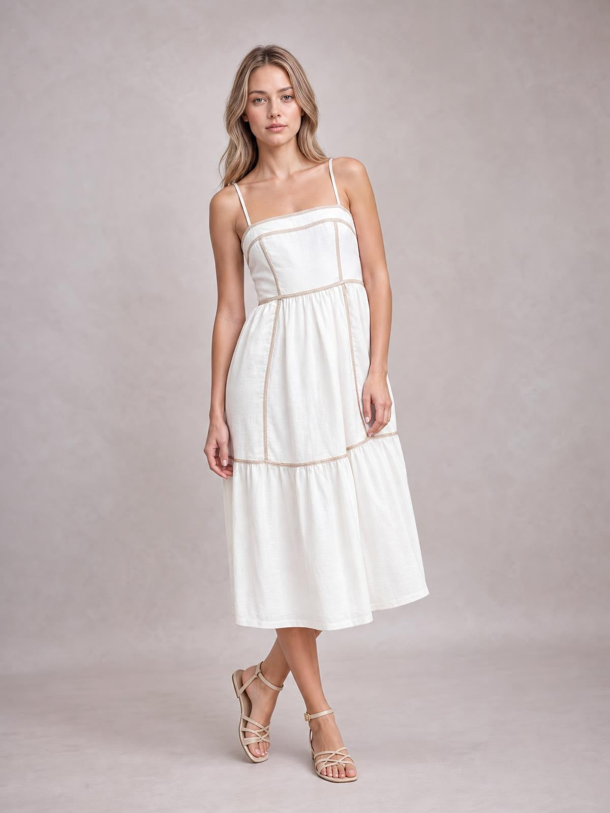 Model poses with crossed legs in a white square-neck midi cami dress featuring thin straps, contrast trim, a defined waist, and a softly tiered silhouette.