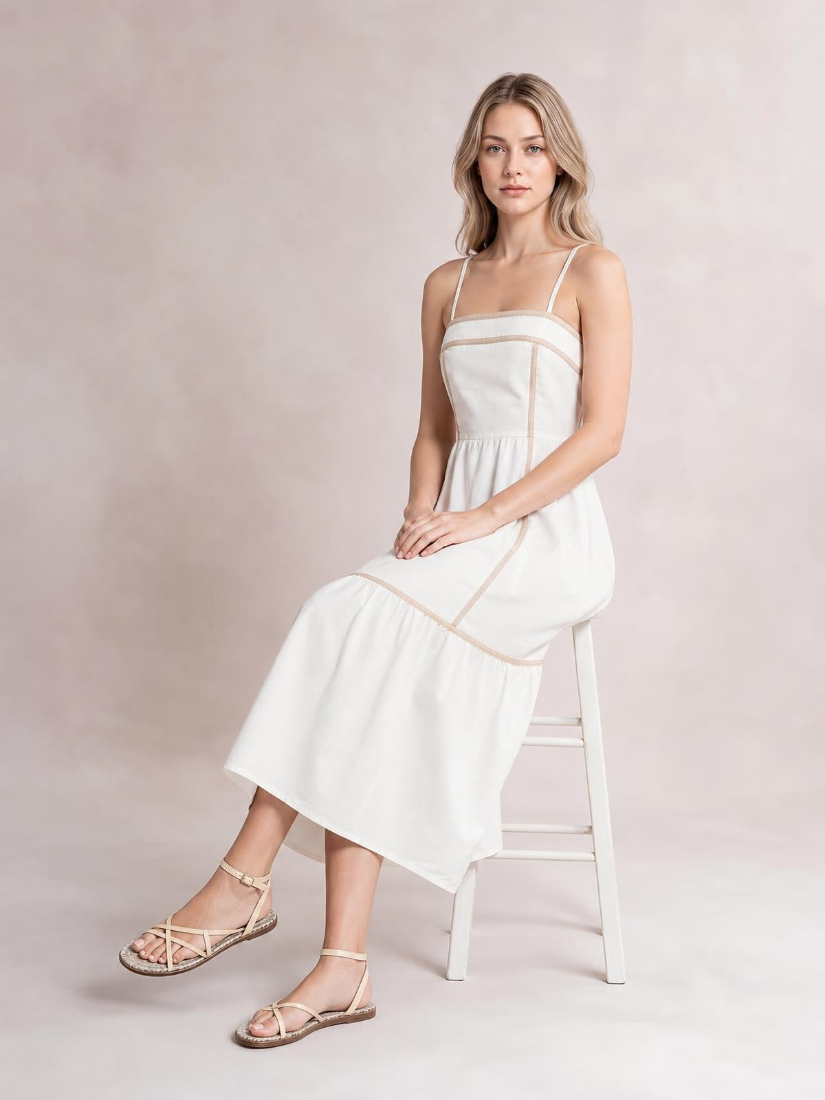 Model seated in a white contrast-trim square-neck midi cami dress with thin straps, a fitted ruched bodice, defined waist, and tiered flowing skirt.