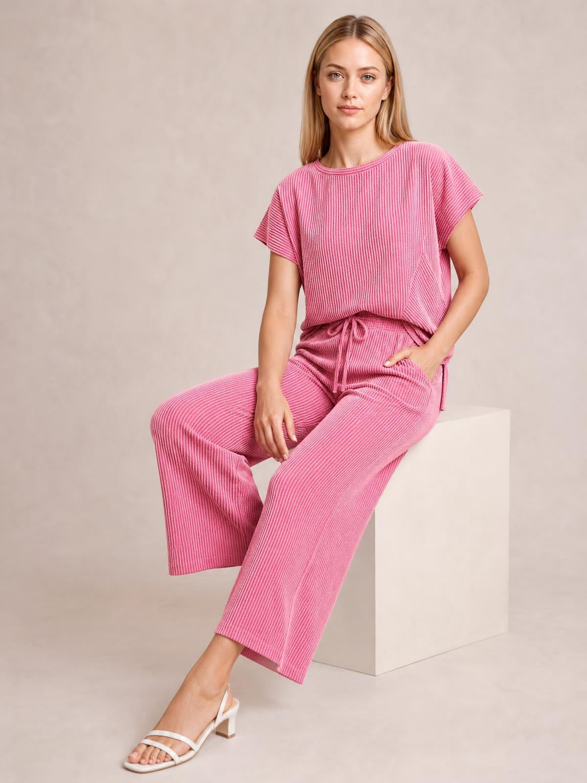 A seated model wears a pink corded knit set with a short-sleeve T-shirt, drawstring waist, and relaxed wide-leg pants.