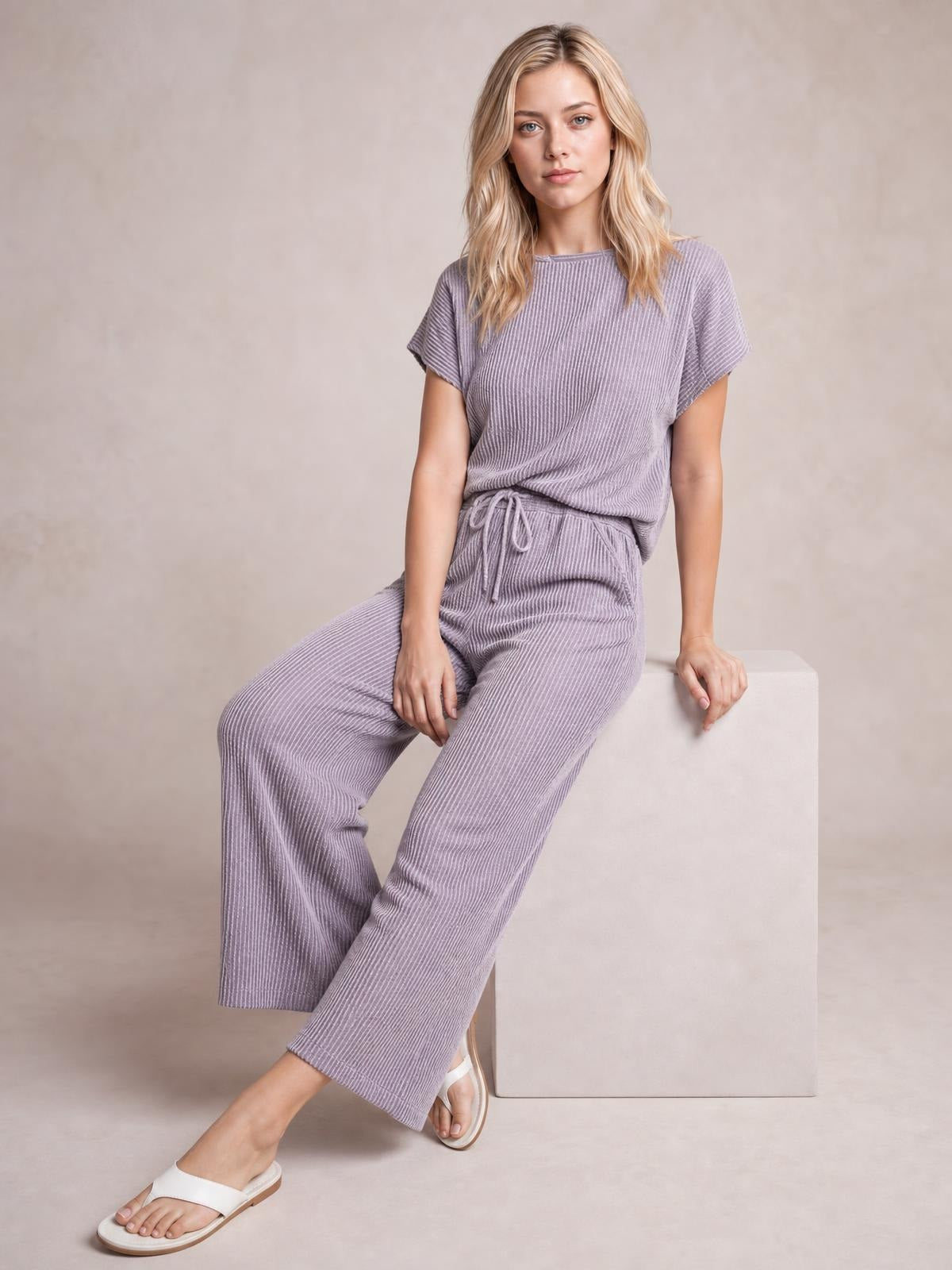 A model sits with crossed legs in a lavender corded knit set featuring a short-sleeve T-shirt and relaxed wide-leg pants with a drawstring waist.
