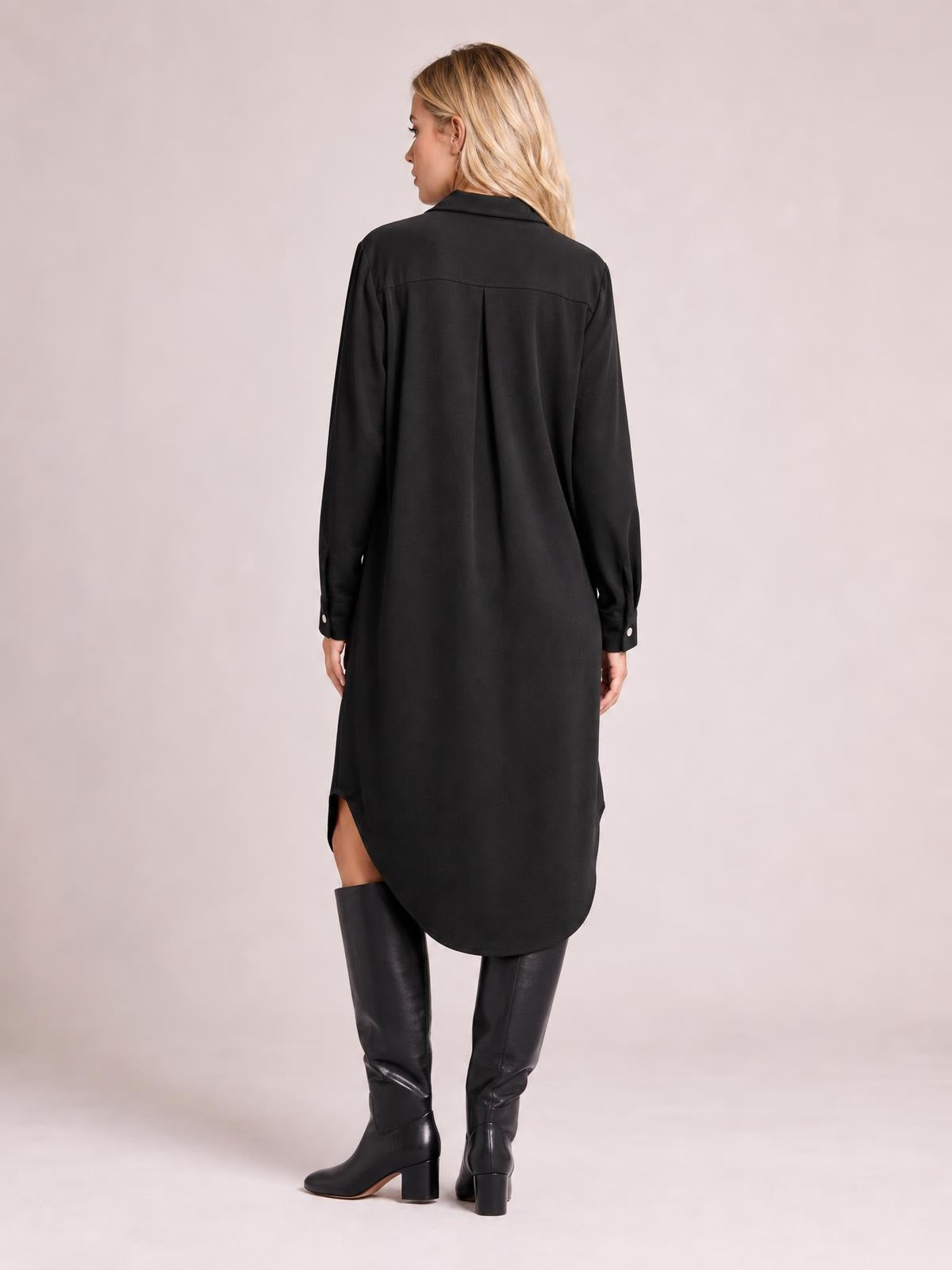 Model shown from the back in a black long-sleeve shirt dress with a collared neckline, relaxed silhouette, back pleat, and rounded shirttail hem.